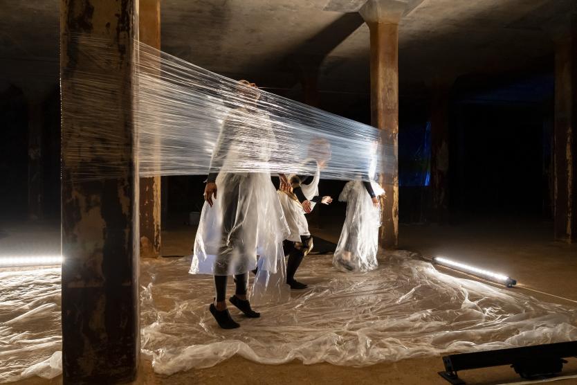Das Bild zeigt mehrere Personen in weißen, transparenten Gewändern, die hinter gespannter Folie zwischen Betonsäulen stehen und von warmem sowie kaltem Licht angestrahlt werden.