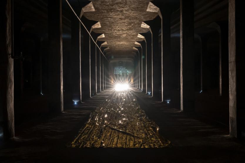 Das Bild zeigt langen Säulengang in dunklem Raum, mit goldglänzendem Folienstreifen am Boden, starkem Lichtschein im Hintergrund und projiziertem Text an der Wand.