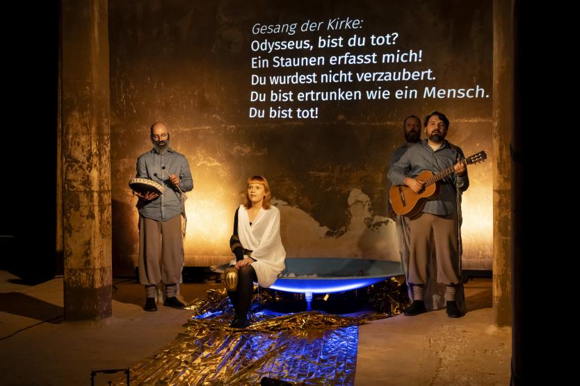 Das Bild zeigt vier Personen in Bühneninszenierung vor projiziertem Text an der Wand, mit einer sitzenden Figur in weißem Gewand auf einer beleuchteten Wanne und drei stehenden Musikern mit Trommel und Gitarre.