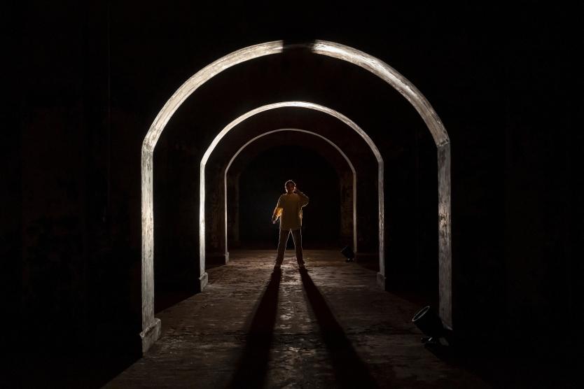 Das Bild zeigt eine Person in hellem Oberteil, stehend unter beleuchtetem Rundbogen in dunklem Raum, mit langen Schatten, die nach vorne auf den Boden fallen.