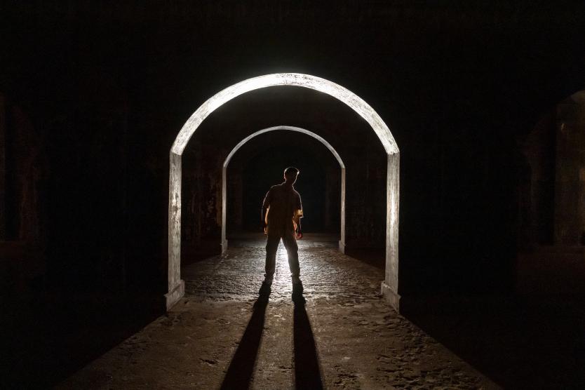 Das Bild zeigt eine einzelne Person in hellem Oberteil, stehend unter beleuchtetem Rundbogen in dunklem Raum, mit langem Schatten, der vom rückwärtigen Licht nach vorne auf den Boden fällt.