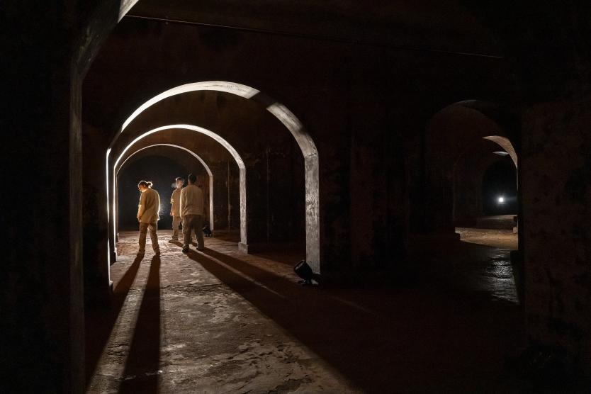 Das Bild zeigt drei Personen in hellen Oberteilen, stehend unter beleuchtetem Rundbogen in dunklem Raum, mit langen Schatten, die von rückwärtigem Licht auf den Boden projiziert werden.
