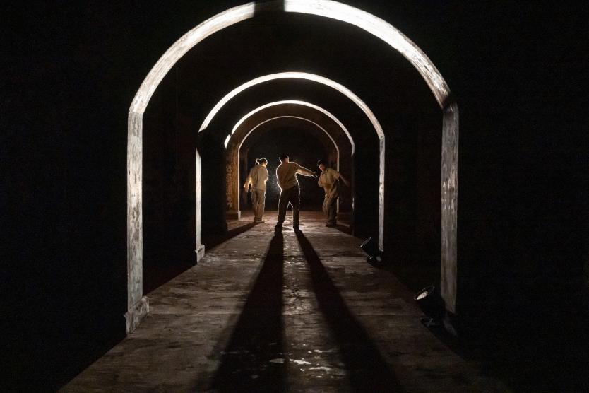 Das Bild zeigt drei Personen in hellen Oberteilen, stehend und bewegend unter hintereinander angeordneten beleuchteten Rundbögen, mit langen Schatten, die nach vorne auf den Boden fallen.